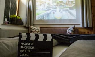 A cozy room with a film clapboard in the foreground and a projector image in the background. The scene features a beautiful outdoor area with water and trees.