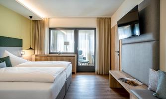 A modern hotel room with two beds and a large window. The furnishings are elegant and bright, with a television and a cozy seating area.