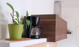 A modern kitchen with a black coffee machine and a green plant on a white shelf. In the background, wooden elements and a hood are visible.