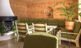 A cozy room with a green seating area and a fireplace nook. The walls are made of light wood, and a pot with a plant is in the background.