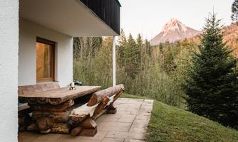 A cozy terrace with a large wooden table, surrounded by trees. In the background, a majestic mountain can be seen.