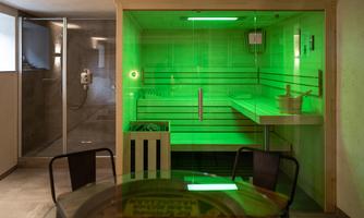 A modern sauna with green lighting and wooden panels. Next to it is a table and a shower in the background.