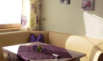 A cozy dining area with a bright corner bench and a table. The table is decorated with a purple tablecloth and surrounded by beige chairs.