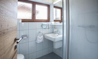 A modern bathroom with a shower, a sink, and windows. The walls are tiled in light colors, and the floor is gray.
