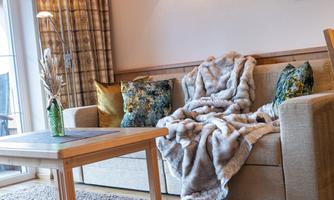 A cozy living room corner with a sofa adorned with a snug blanket and decorative cushions. A small table and a vase with dried plants complement the decor.