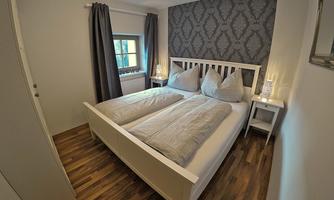 A cozy bedroom with a white bed and gray bedding. The walls are decorated with a modern pattern, and there is a window with curtains.