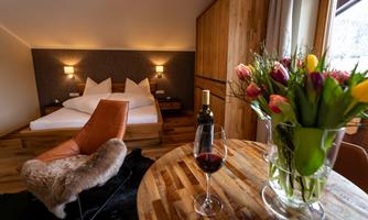 A cozy room with a double bed and warm wooden paneling. On the table are a glass of wine and a bouquet of flowers.