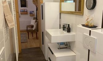 A modern bathroom with white furniture and a large mirror. In the background, there is a hallway with a wooden chair and pictures visible.