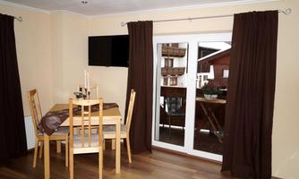 A cozy dining area with a table and chairs. Dark curtains hang in front of a balcony window, and a television is mounted on the wall.