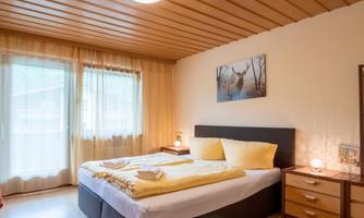 A cozy bedroom with a double bed and yellow pillows. Large windows let in plenty of light and there is an attractive wall decoration.