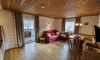 A bright living room with a wooden ceiling and parquet floor. It is furnished with a red sofa, a dining table, and windows with a view.