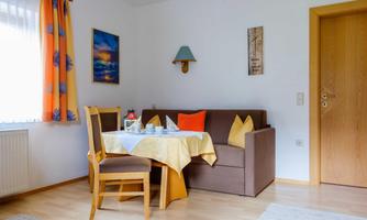 A bright living room with a cozy sofa and a table. Decorative pictures hang on the walls, and the windows are adorned with colorful curtains.