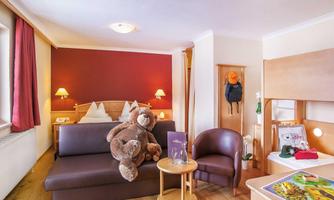 A cozy hotel room with a large bed and a comfortable sofa. A plush bear sits on the sofa, and there is a small seating area with a table.