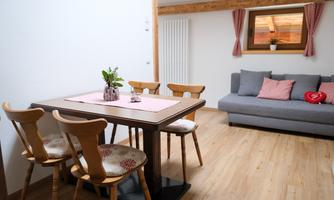 A cozy living space with a dining table and chairs as well as a comfortable sofa. The windows with curtains and the wooden-paneled walls create an inviting atmosphere.