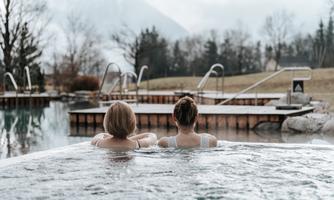 Two people are relaxing in a warm water pool. In the background, there are more pools and a picturesque landscape.