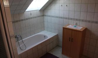 A bright bathroom with a window and bathroom tiles. There is a bathtub and a wooden cabinet for storage.