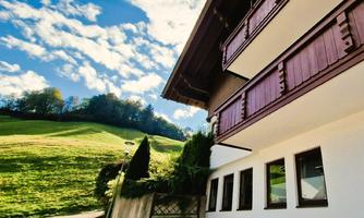 A charming house with a balcony and large windows. In the background, green meadows and a blue sky with clouds can be seen.