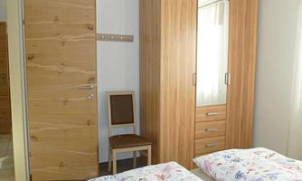 A simple bedroom with two beds, a wooden wardrobe, and a chair. The walls are neutral, and the room appears bright and friendly.