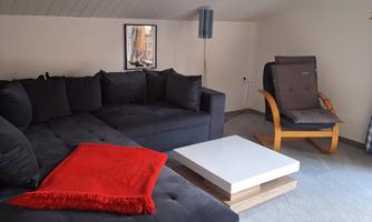 A cozy living room with a dark couch and a white table. A red plaid is draped over the couch, while a comfortable armchair stands beside it.
