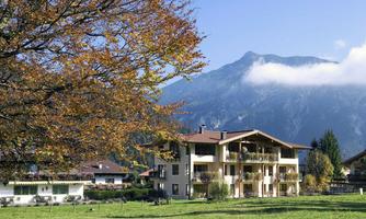 A picturesque house with multiple floors and large windows, surrounded by autumnal trees and an impressive mountain landscape in the background. The sky is clear with a few clouds.