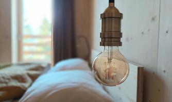 A modern light bulb hangs over a bed in a cozy room. In the background, windows and curtains can be seen, allowing light in.