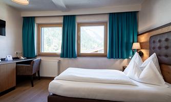 A modern hotel room with a double bed, a desk, and a cozy chair. The large windows offer a view of the mountains and let in plenty of daylight.