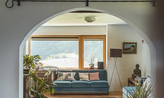 A cozy living room with a blue sofa and large windows that let in daylight. The room is decorated with plants and a colorful rug.