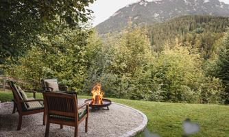A cozy seating area with wooden furniture and a campfire. In the background, green mountains and trees stretch out.