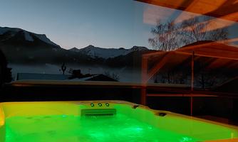 A hot tub with green lighting is in the foreground. In the background, mountains are visible under a clear sky.