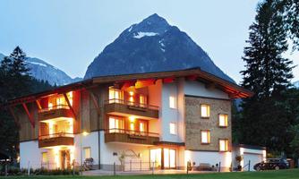 A modern house with wooden cladding, surrounded by an impressive mountain. The warm lights of the building shine in the dusk.