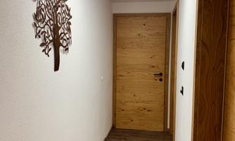 A bright corridor with plain walls and a wooden floor. A decorative tree sculpture hangs on the wall, and there is a modern ceiling light.