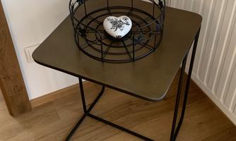 A stylish side table made of metal with an oval top and a black decorative basket on it. The basket contains a heart-shaped object in the middle.