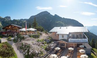A mountain inn with a sun terrace and outdoor tables. In the background, majestic mountains and a clear blue sky are visible.