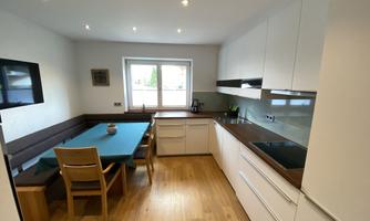 A modern kitchen with white furniture and wooden flooring. A dining table with chairs is placed next to a window.