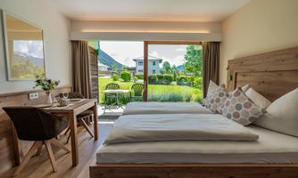 A cozy bedroom with a large bed and a wooden desk. Through the window, there is a beautiful view of a garden and comfortable chairs.
