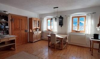 A cozy room with wooden floors and large windows. There is a table with chairs, as well as a wardrobe and a pendant lamp.