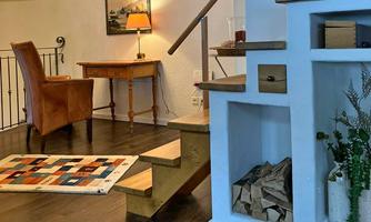 A cozy living space with an armchair, a small table, and a wooden staircase. The room is bright and cheerful, with a carpet and a firewood storage.