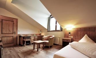 A cozy room with slanted walls and wooden details. There is a bed, a table, and a chair, as well as a window with a view outside.