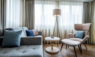 A modern living room with a gray sofa, a beige armchair, and a small table. Large windows with curtains allow plenty of daylight to enter.