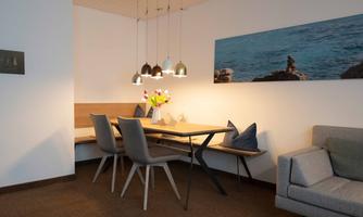 A cozy dining area with a wooden table, chairs, and a bench. Fresh flowers are on the table, and soft light creates an inviting atmosphere.