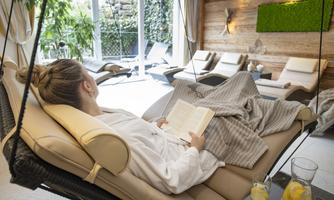 A relaxed person in a bathrobe is lying in a hammock and reading a book. In the background, comfortable loungers and green plants can be seen.