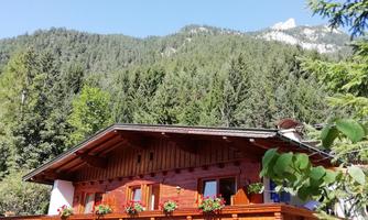 A traditional chalet with a wooden balcony, surrounded by green trees. Majestic mountains are visible in the background.