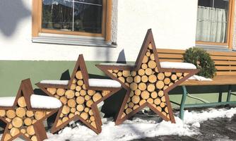 Three decorative wooden stars stand on a snowy surface in front of a house. The stars are made from wooden logs and have a rustic appearance.