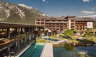 A modern hotel with beautiful architecture and large windows. In the foreground, pools and a picturesque surroundings with mountains can be seen.