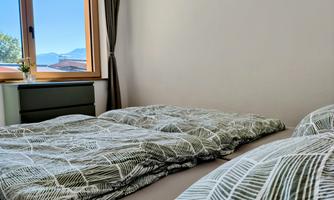 A cozy bedroom with two beds and bedding in a modern leaf pattern. Soft light streams through the window, and trees and mountains are visible in the background.
