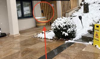 A front door with a key box in the foreground. The ground is covered with snow and plants are visible in the background.