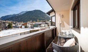 A balcony with a table and chairs offers a view of snow-covered mountains and a picturesque village. The sky is clear and the landscape is idyllic.