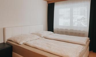 A bright bedroom with a cozy double bed and white bed linens. Large windows with curtains allow for plenty of daylight.
