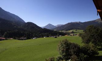 A vast, green meadow surrounded by mountains under a clear blue sky. In the background, small villages and forests are visible.