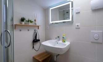 A modern bathroom with a sink, a lit mirror, and a simple wooden shelf. Plants and care products create a cozy atmosphere.